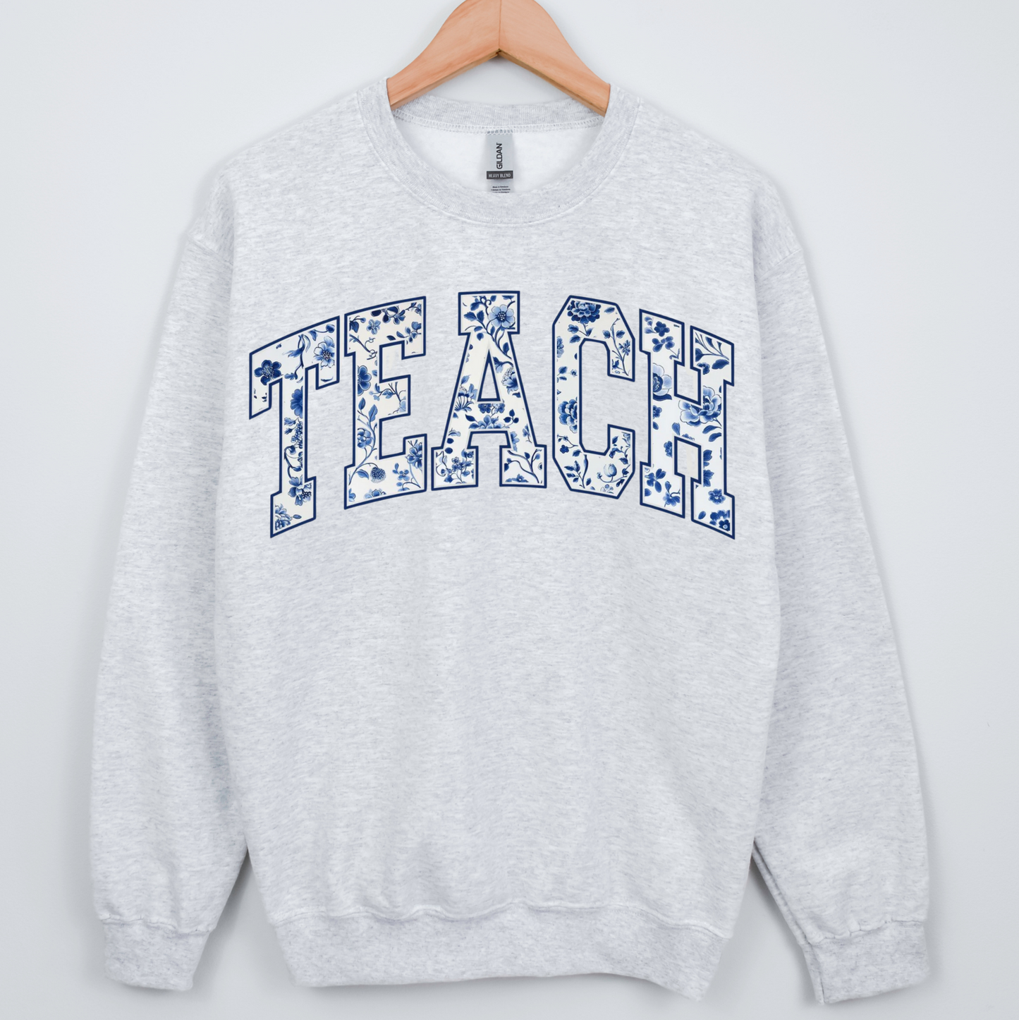 Gray sweatshirt with 'TEACH' printed on it, hanging on a wooden hanger against a white background.
